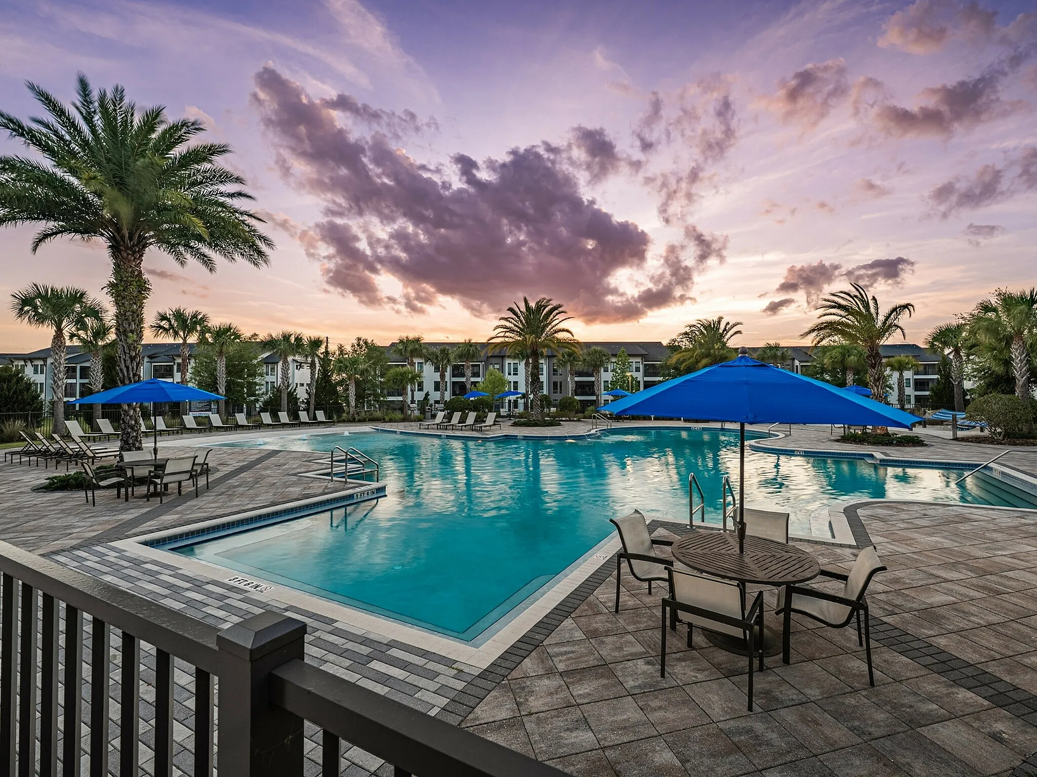 Luxurious resort-style swimming pool with palm trees, comfortable lounge chairs, and stylish blue umbrellas at sunset.