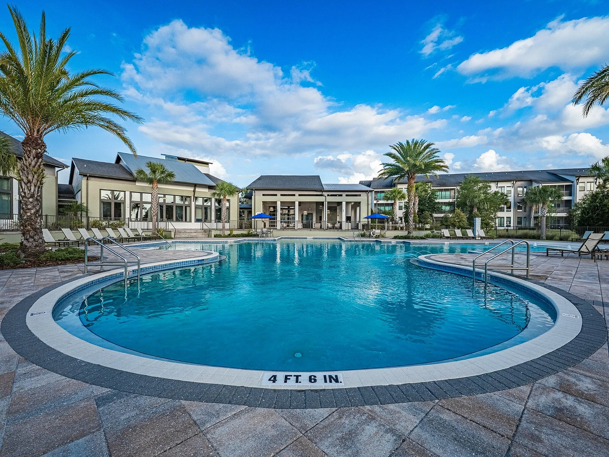 Luxurious apartment complex with inviting round outdoor pool, surrounded by palm trees and modern lounge chairs, set against a bright blue sky.