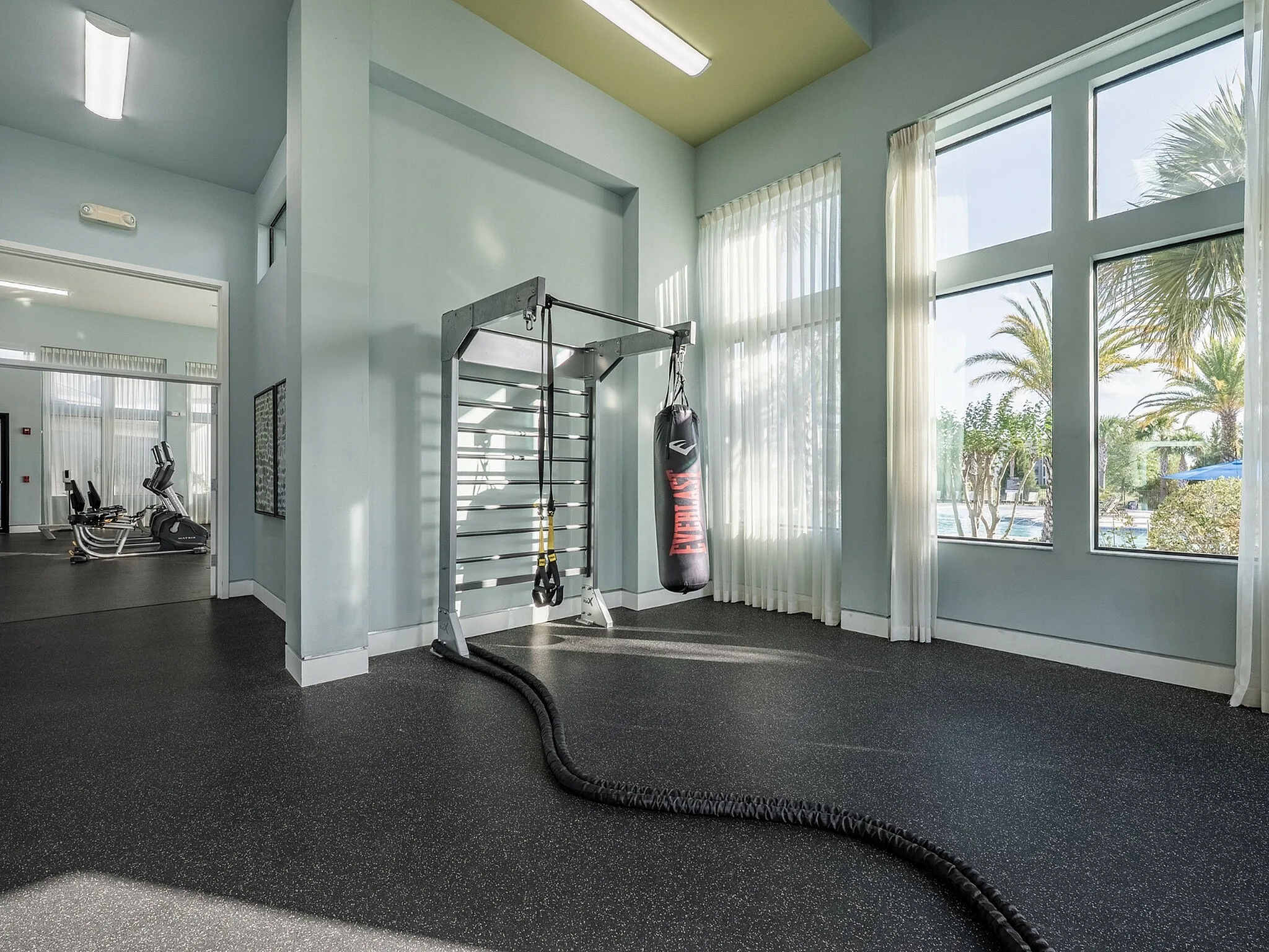 Spacious fitness area with modern gym equipment, boxing setup, and large windows offering a refreshing view of palm trees and a pool.
