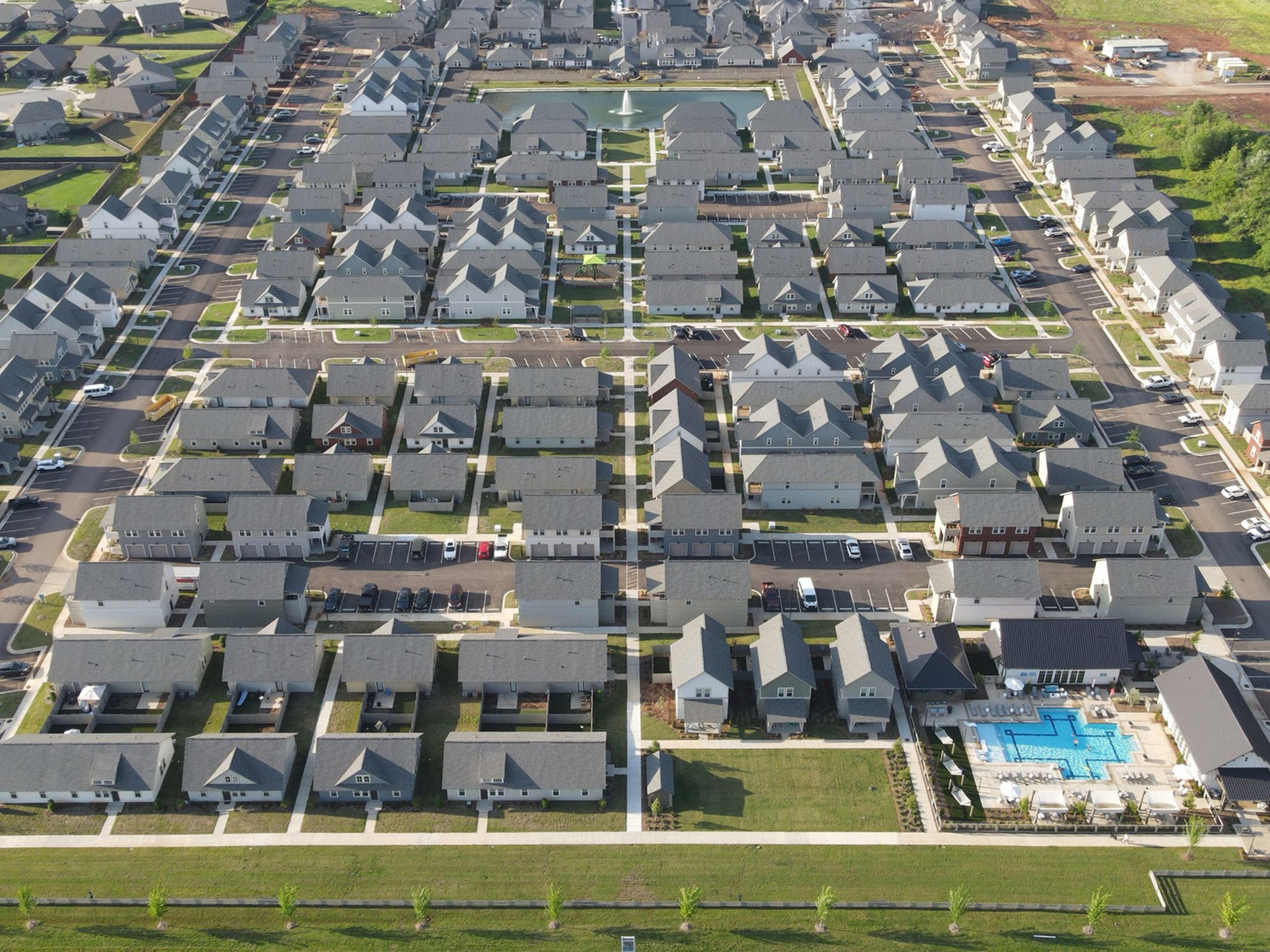 Aerial view of a modern residential community showcasing neatly arranged houses, lush green lawns, and a central swimming pool area, offering a serene suburban lifestyle.