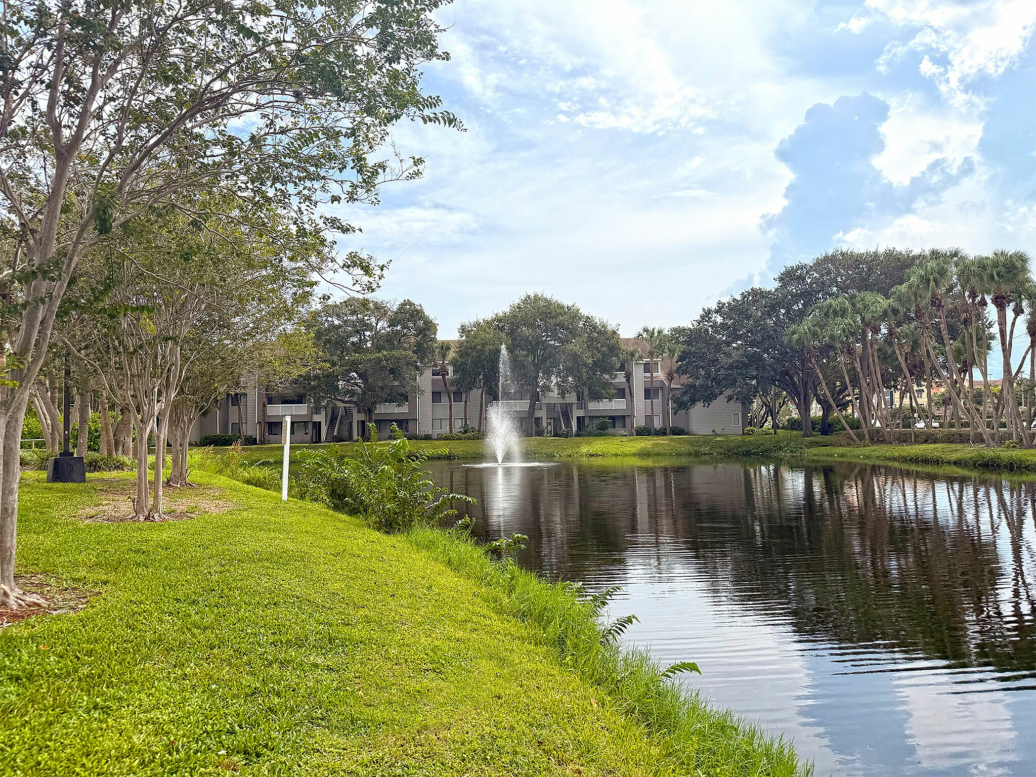 Scenic apartment view with lush greenery, reflecting pond, and elegant fountain, offering serene living in a tranquil setting.