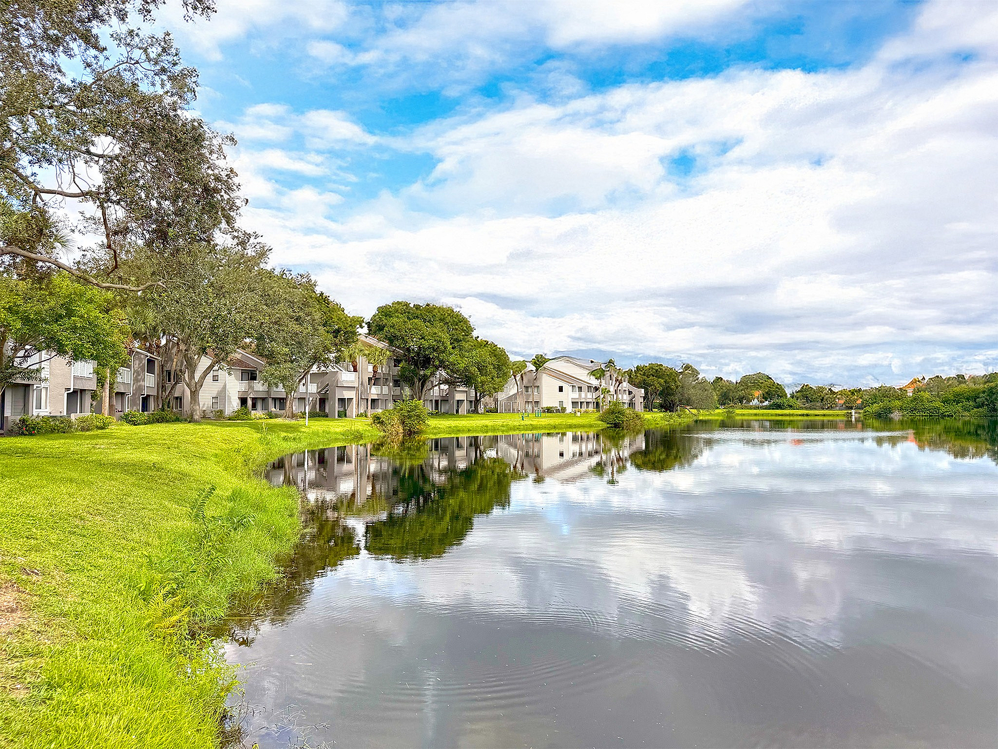 Charming riverside apartments surrounded by lush greenery and tranquil waters, offering serene living with stunning reflections and blue skies.