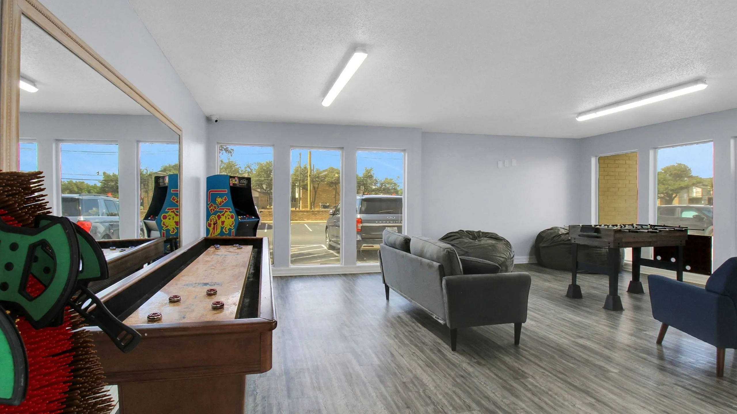 Modern entertainment room with arcade games, shuffleboard, and foosball, featuring comfortable seating and natural light.