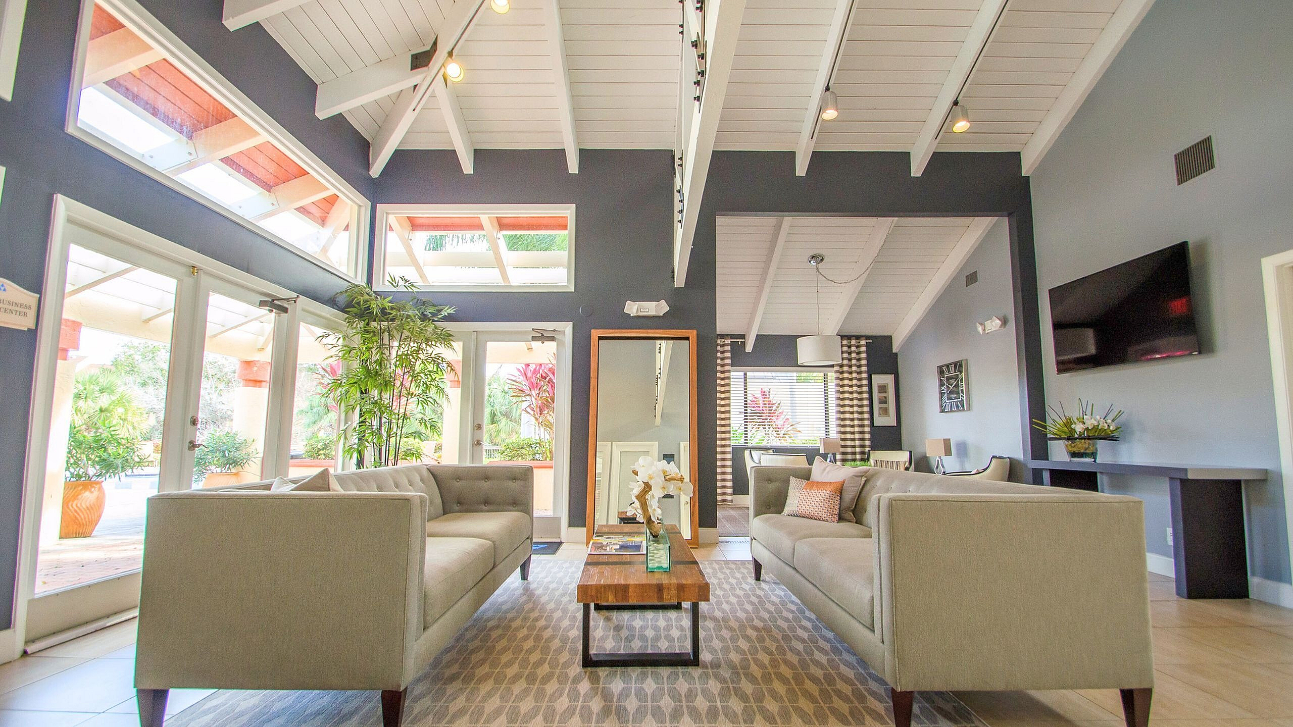 Bright and inviting living room with modern sofas, large windows, high vaulted ceiling, and lush greenery views.