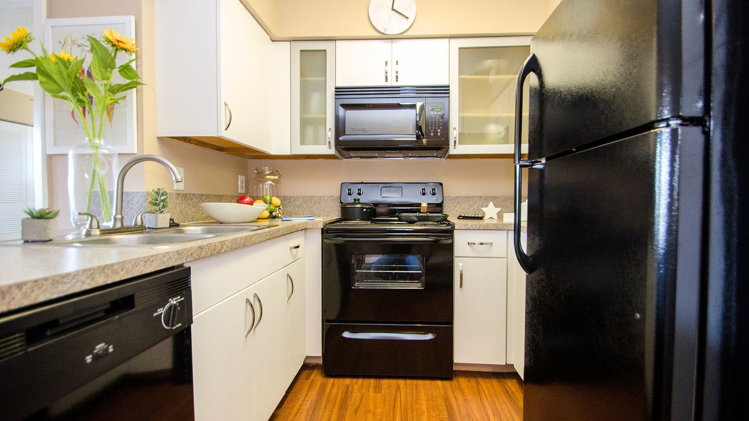 Inviting kitchen with sleek black appliances, modern countertops, and elegant cabinetry, accented by vibrant sunflowers and cozy wooden flooring.