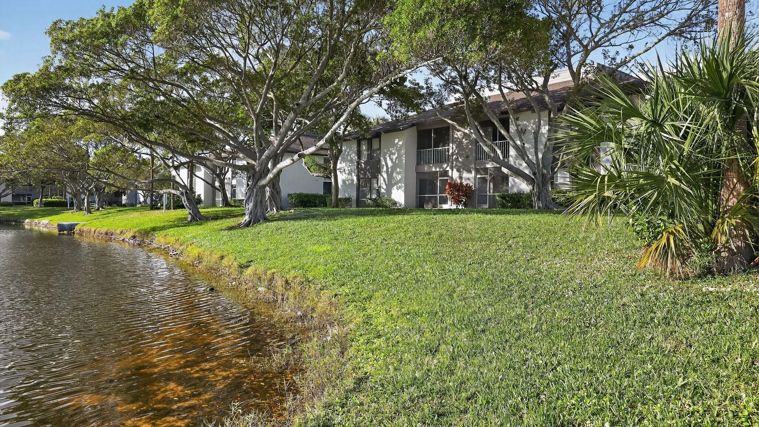 Serene lakeside apartment exterior with lush green lawn and majestic trees creating a tranquil living environment.