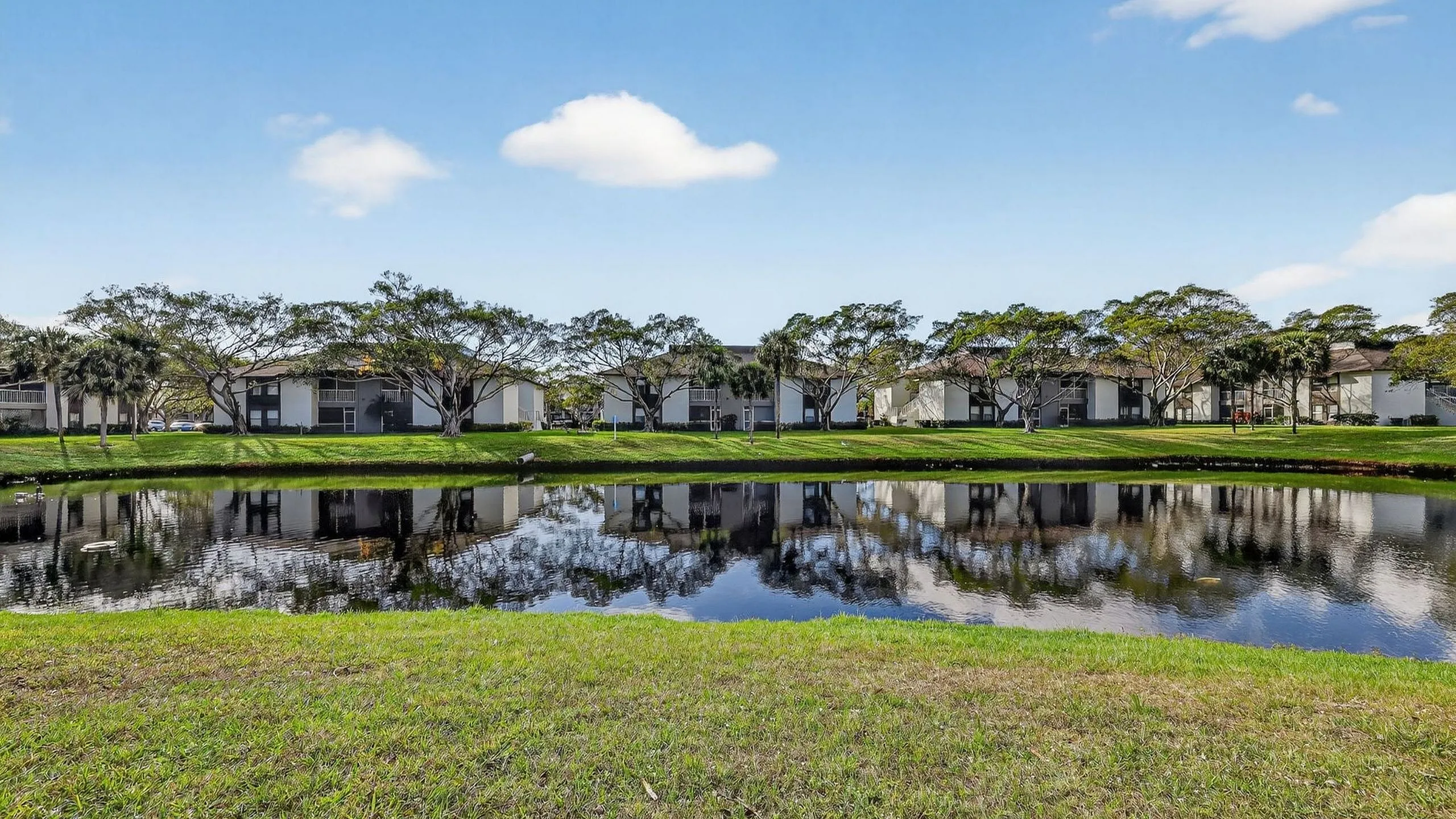 Serene waterfront residences reflecting in a tranquil pond, surrounded by lush greenery and clear blue skies.