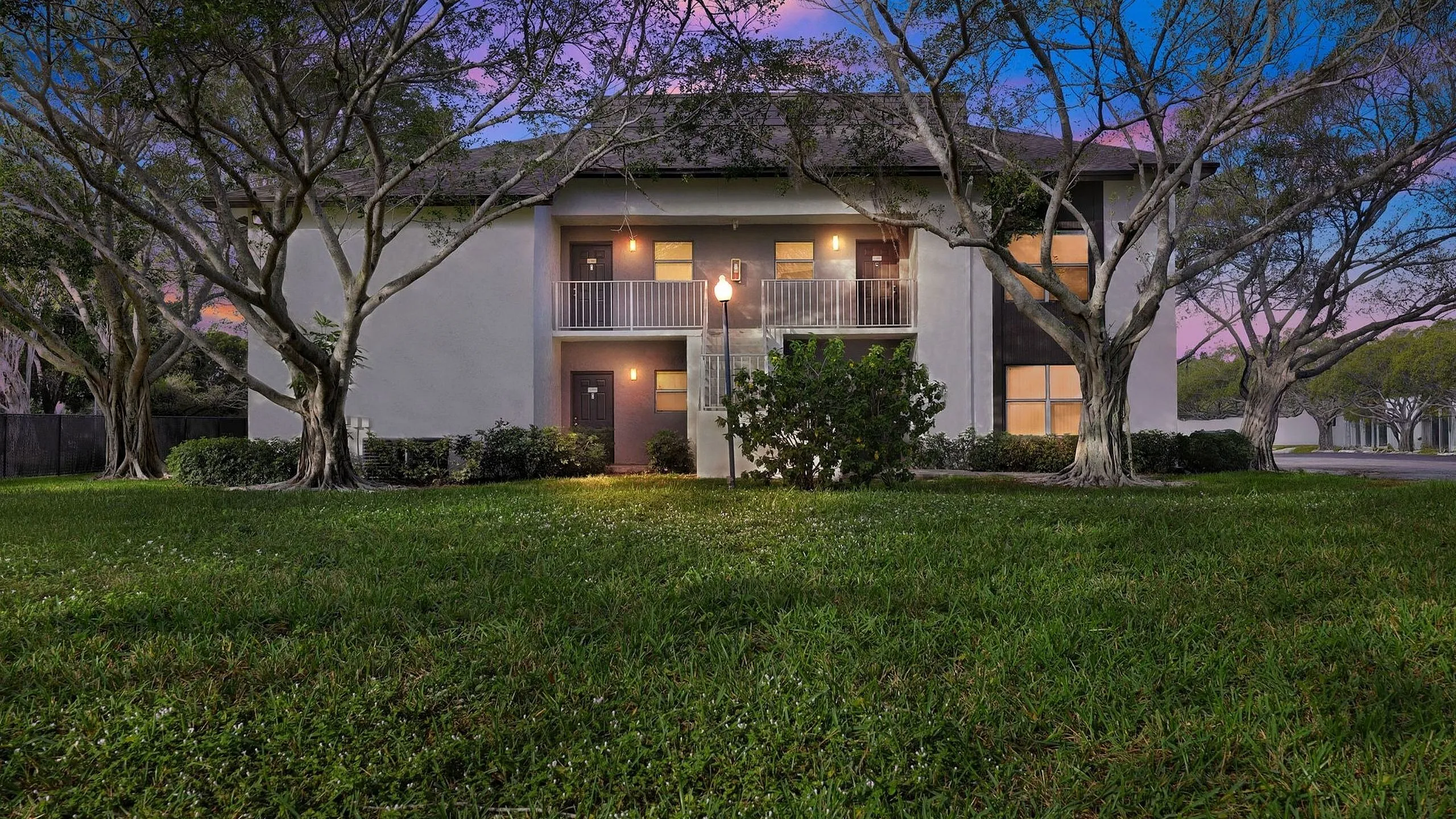 Charming apartment building surrounded by lush greenery and mature trees at dusk, offering serene living with inviting balconies and well-lit entrances.