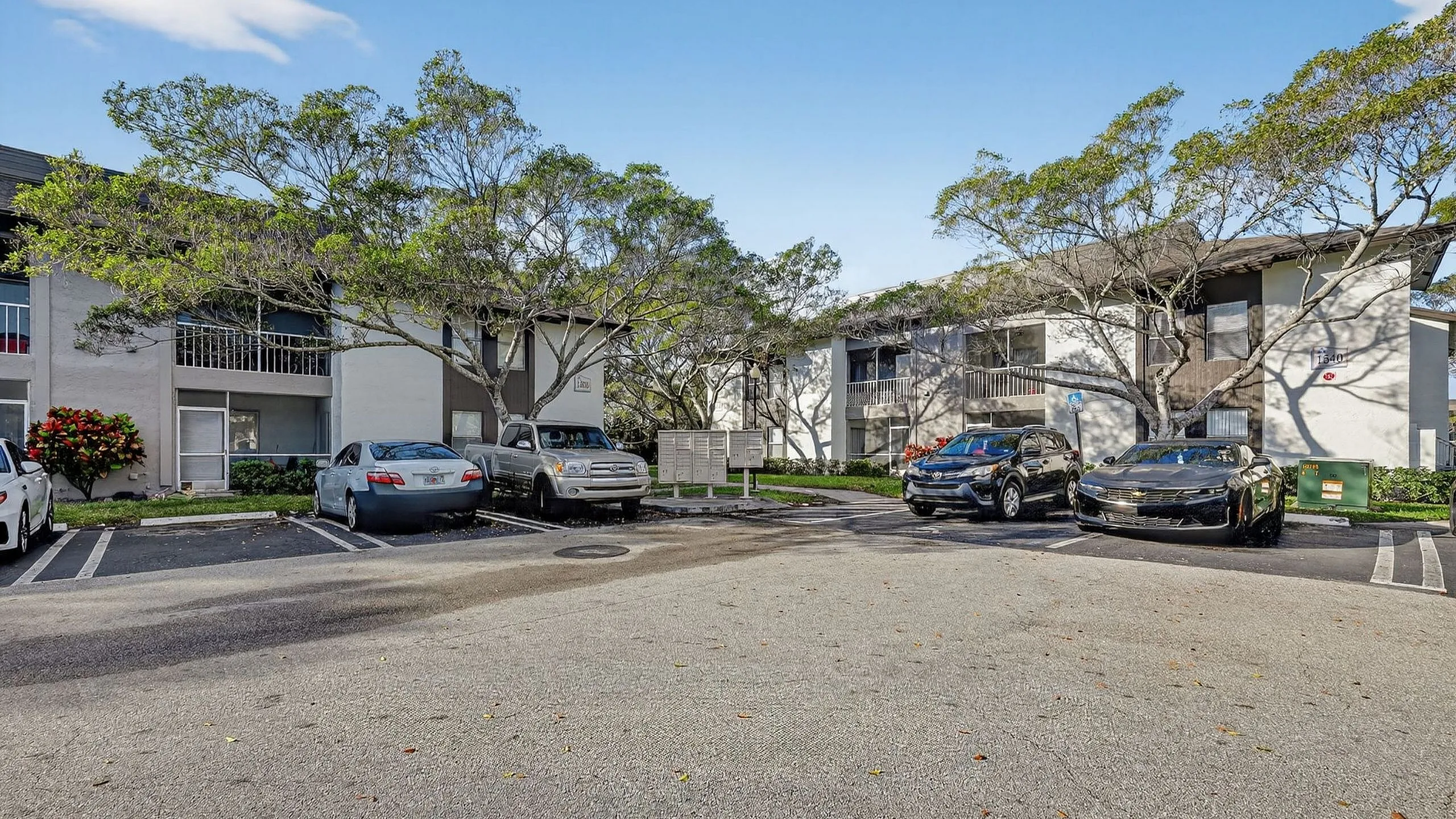 Charming apartment complex with lush trees and ample parking, offering serene living under a bright blue sky.