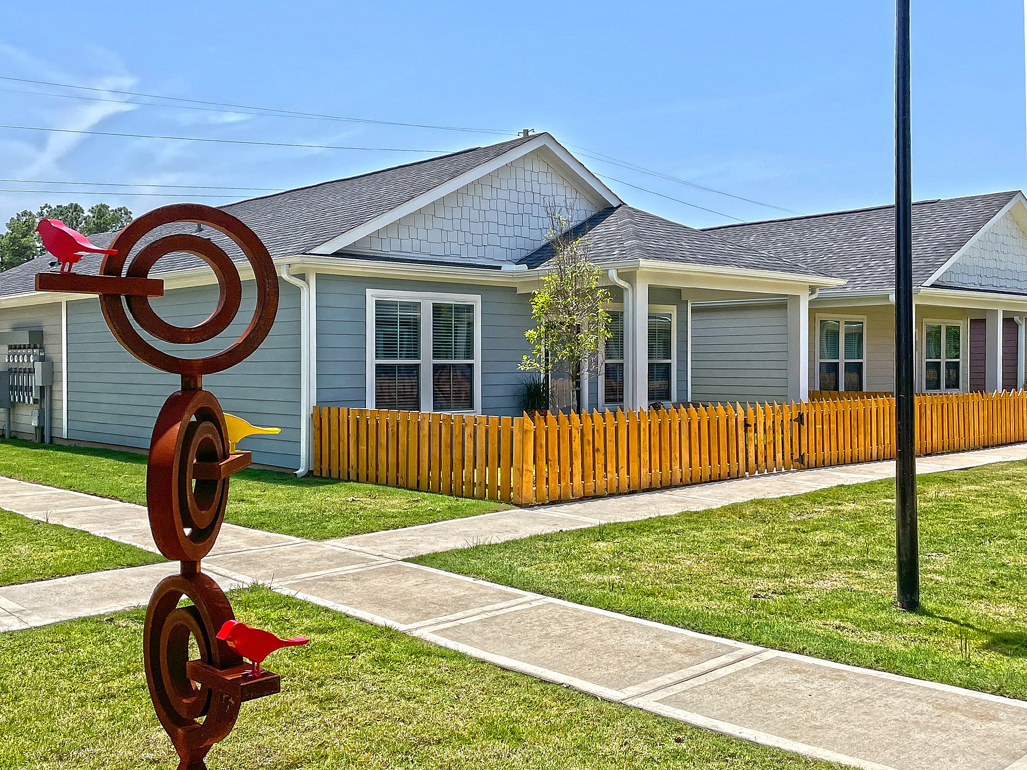 Charming single-story home with pristine gray siding, cheerful yellow fence, and bold artistic sculpture in a lush green setting.