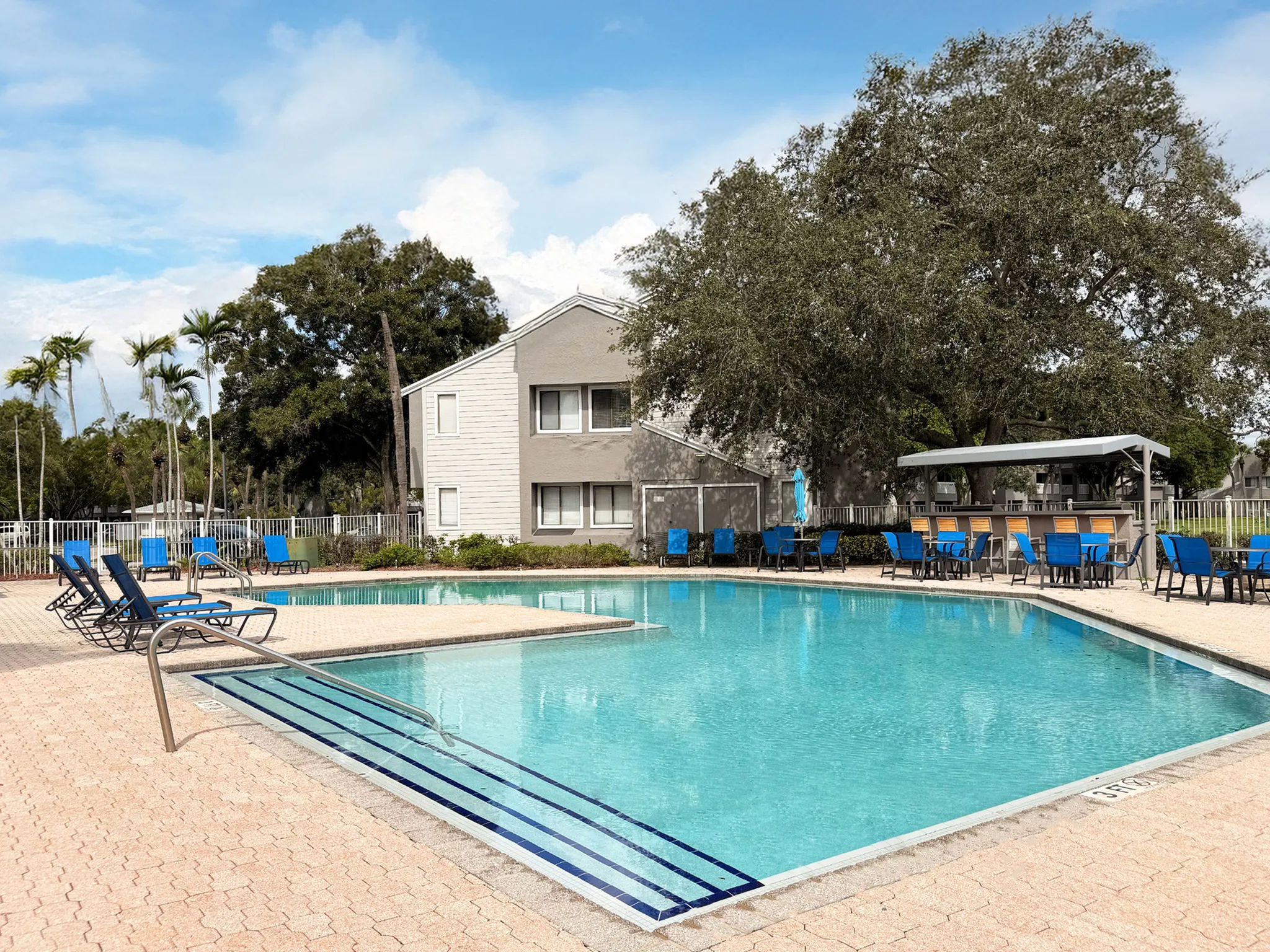 Charming apartment community with a sparkling swimming pool surrounded by blue lounge chairs and lush greenery under a bright sky.