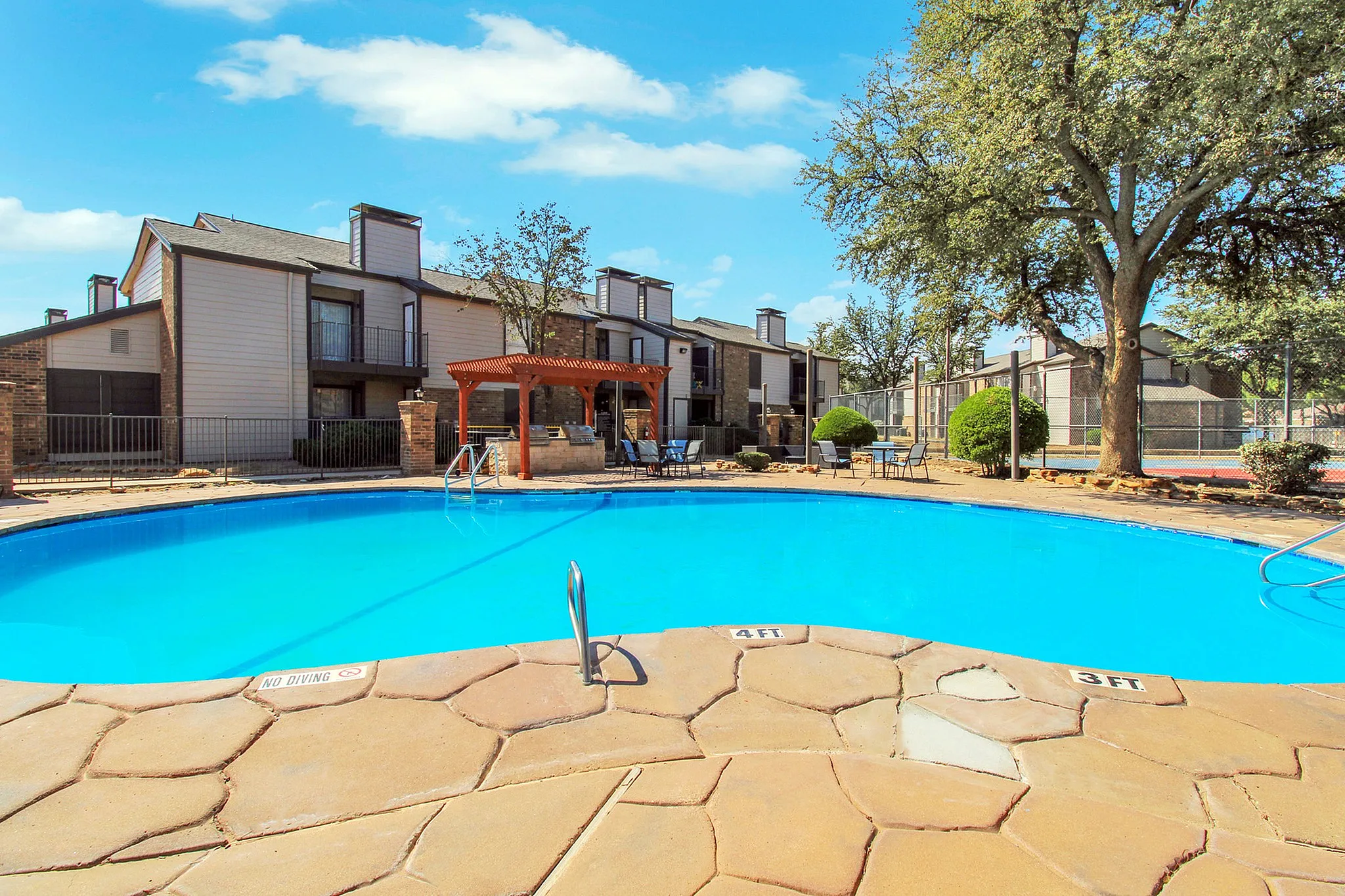 Inviting apartment complex with a sparkling blue swimming pool, surrounded by lush greenery and a charming wooden pergola, perfect for relaxation and outdoor gatherings.