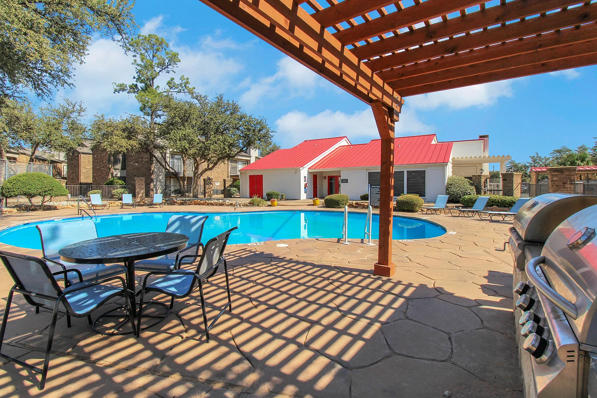 Inviting outdoor pool area with modern seating, surrounded by lush greenery and charming red-roofed clubhouse, perfect for relaxation and leisure.