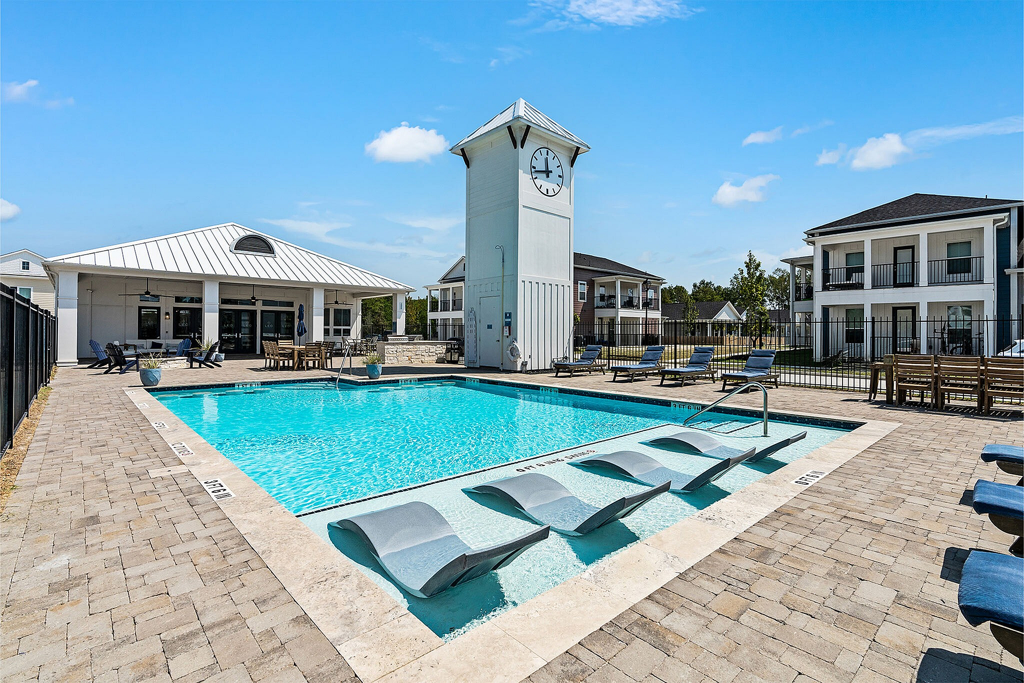 Luxurious apartment complex featuring a sparkling pool, comfortable lounge chairs, and a stylish clock tower under a clear blue sky.