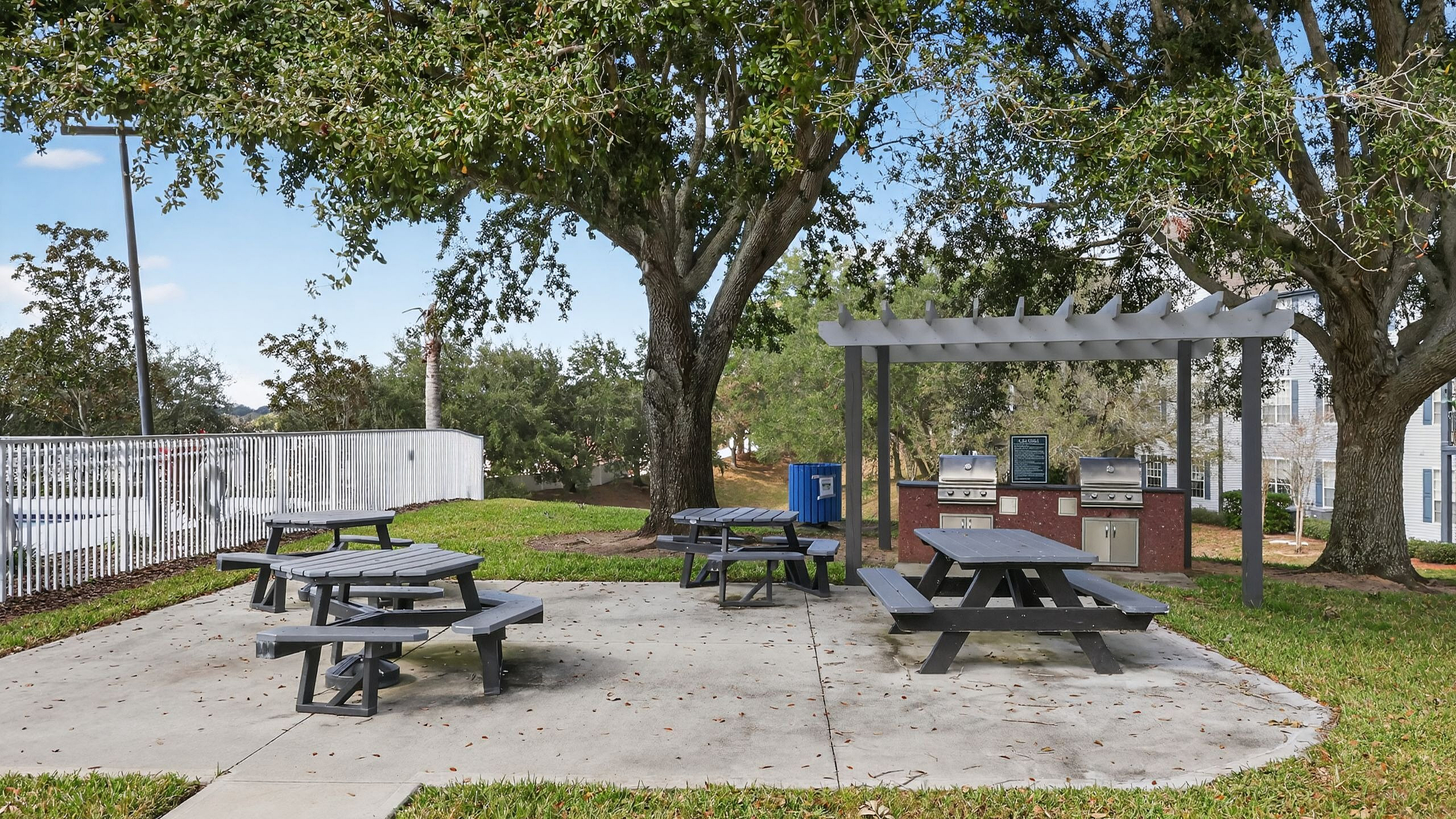 Inviting outdoor picnic area with multiple picnic tables under lush trees and a modern BBQ grill station, perfect for gatherings.