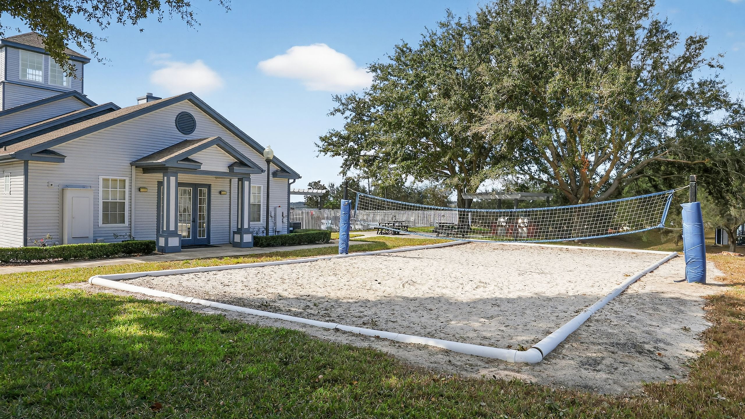 Charming residential clubhouse with adjacent sand volleyball court, set against a backdrop of lush greenery and bright blue skies.