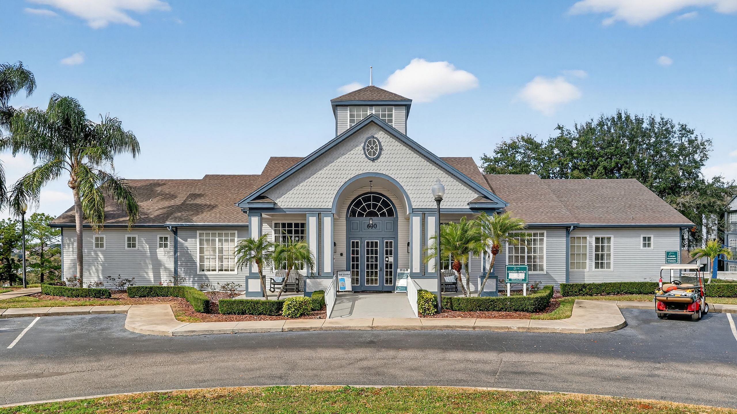 Charming clubhouse with classic architecture, surrounded by lush landscaping and palm trees, offering an inviting entrance and ample parking.