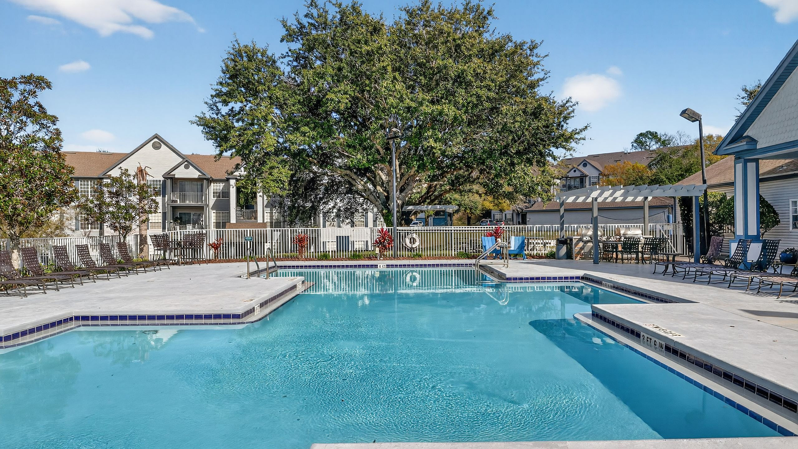 Inviting outdoor pool area with sun loungers, surrounded by lush landscaping and modern apartment buildings under a clear blue sky.