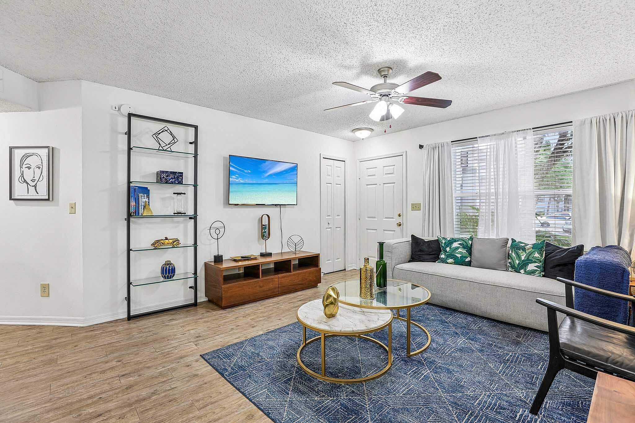 Inviting living room with modern decor, featuring a stylish sofa, elegant glass coffee tables, and a wall-mounted flat-screen TV, all bathed in natural light from large windows.