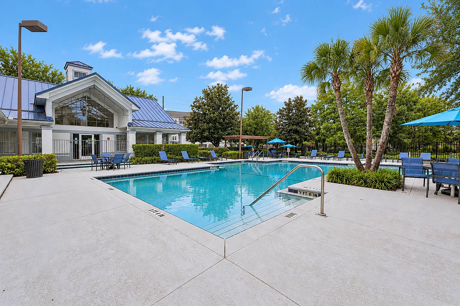 Inviting apartment pool area featuring crystal-clear water, elegant poolside seating, and lush landscaping under a bright blue sky.