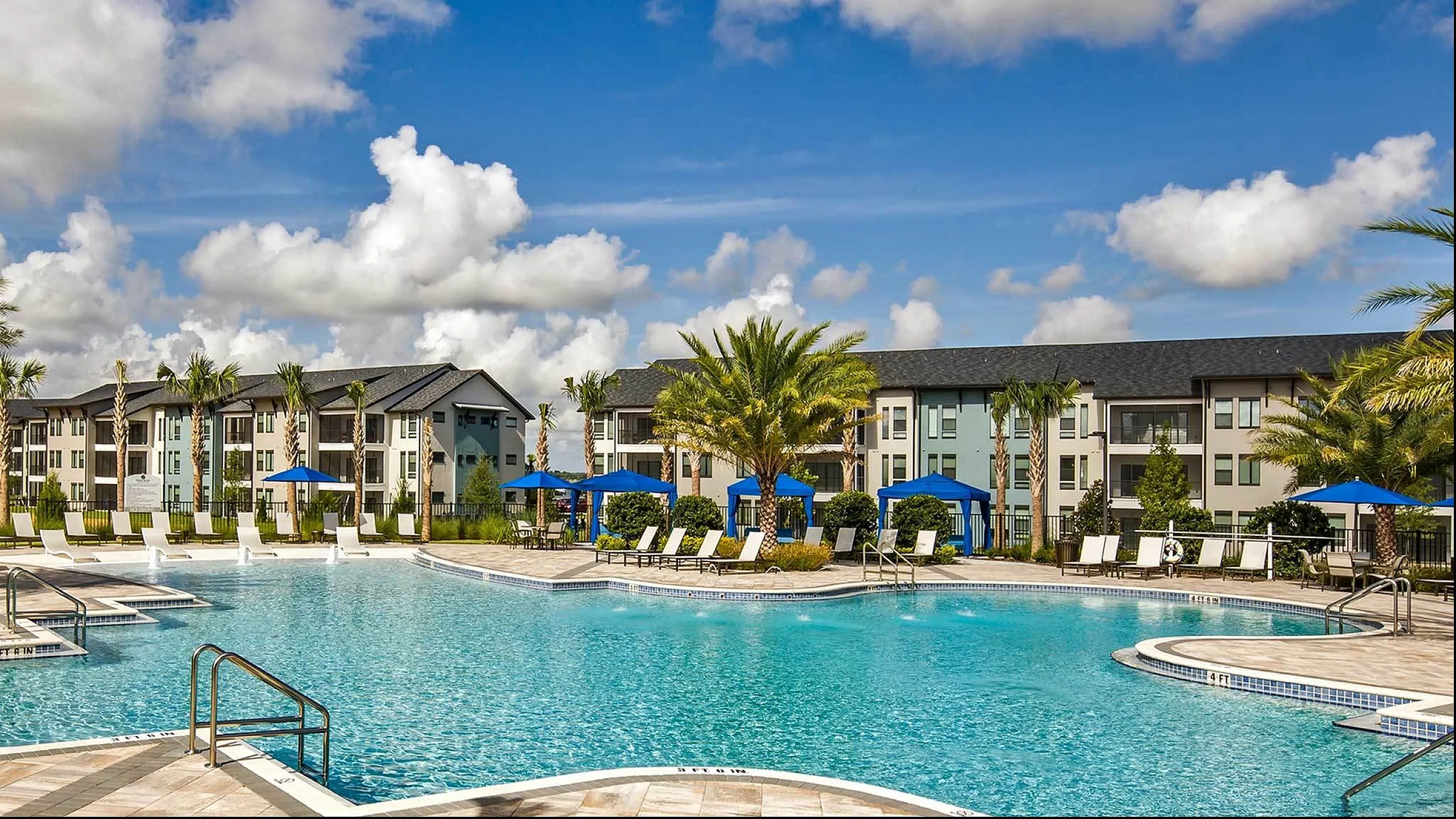 Spacious pool area with sun loungers and umbrellas at a modern apartment complex under a blue sky with fluffy clouds.