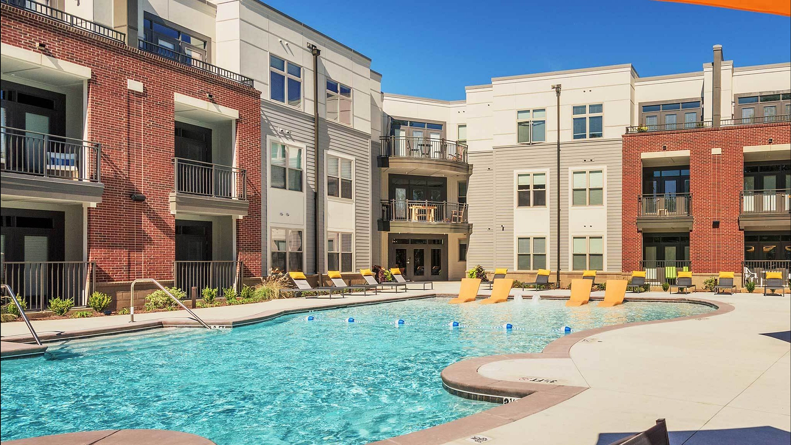 Modern apartment complex with sparkling swimming pool and lounge chairs in a sunlit courtyard.