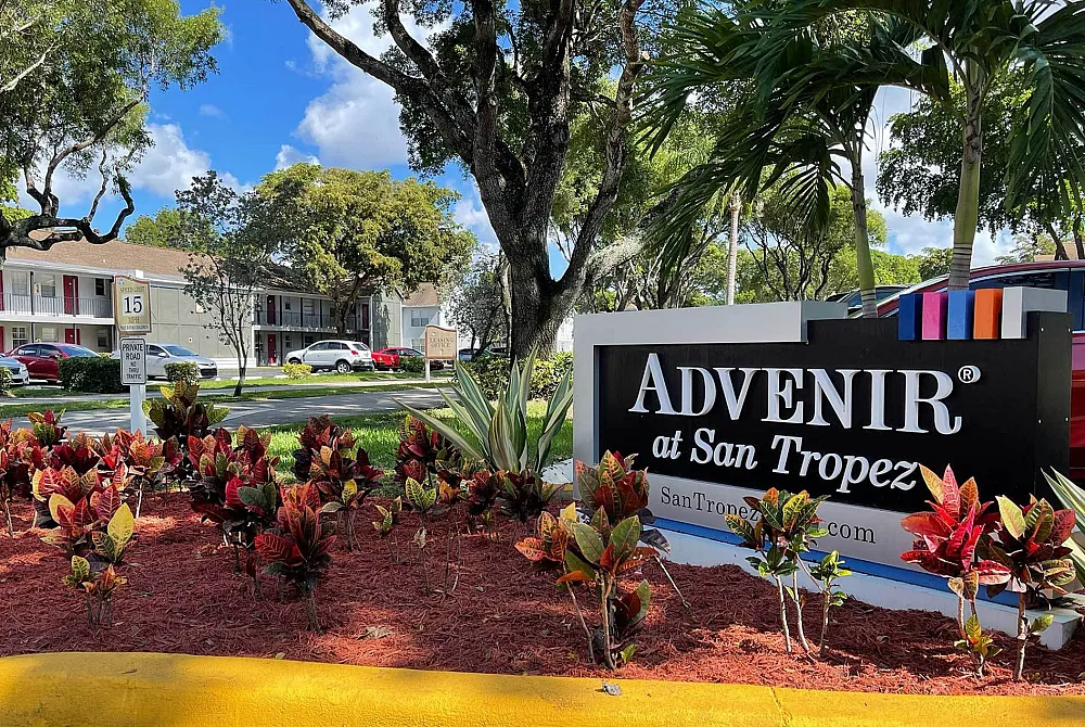 Lush landscaping and vibrant signage at Advenir at San Tropez apartments under a clear blue sky.