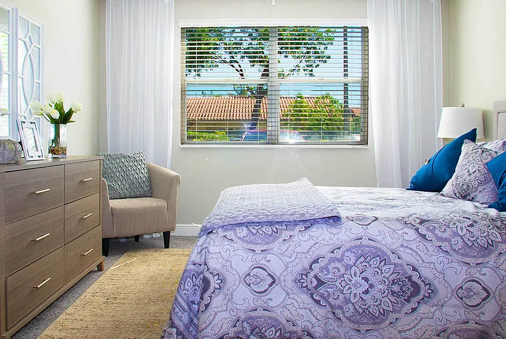 Bright and airy bedroom with elegant decor, featuring a soft pattern bedspread, cozy armchair, and a sunlit window view.