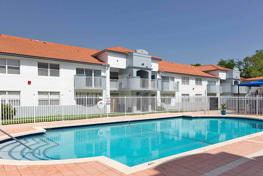 Modern apartment complex with a spacious swimming pool and elegant white fencing under a clear blue sky.