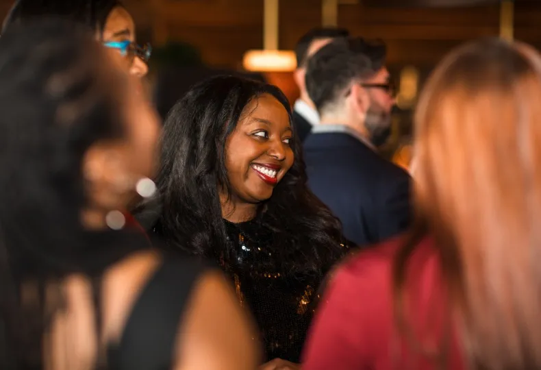 A woman in a black outfit smiling joyfully at a lively social gathering.