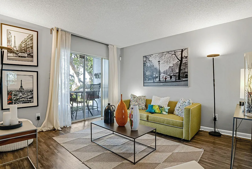 Chic living room with a modern green sofa, vibrant cushions, and a stylish coffee table. Sunlit space with floor-to-ceiling windows opening to a cozy balcony, perfect for relaxation.