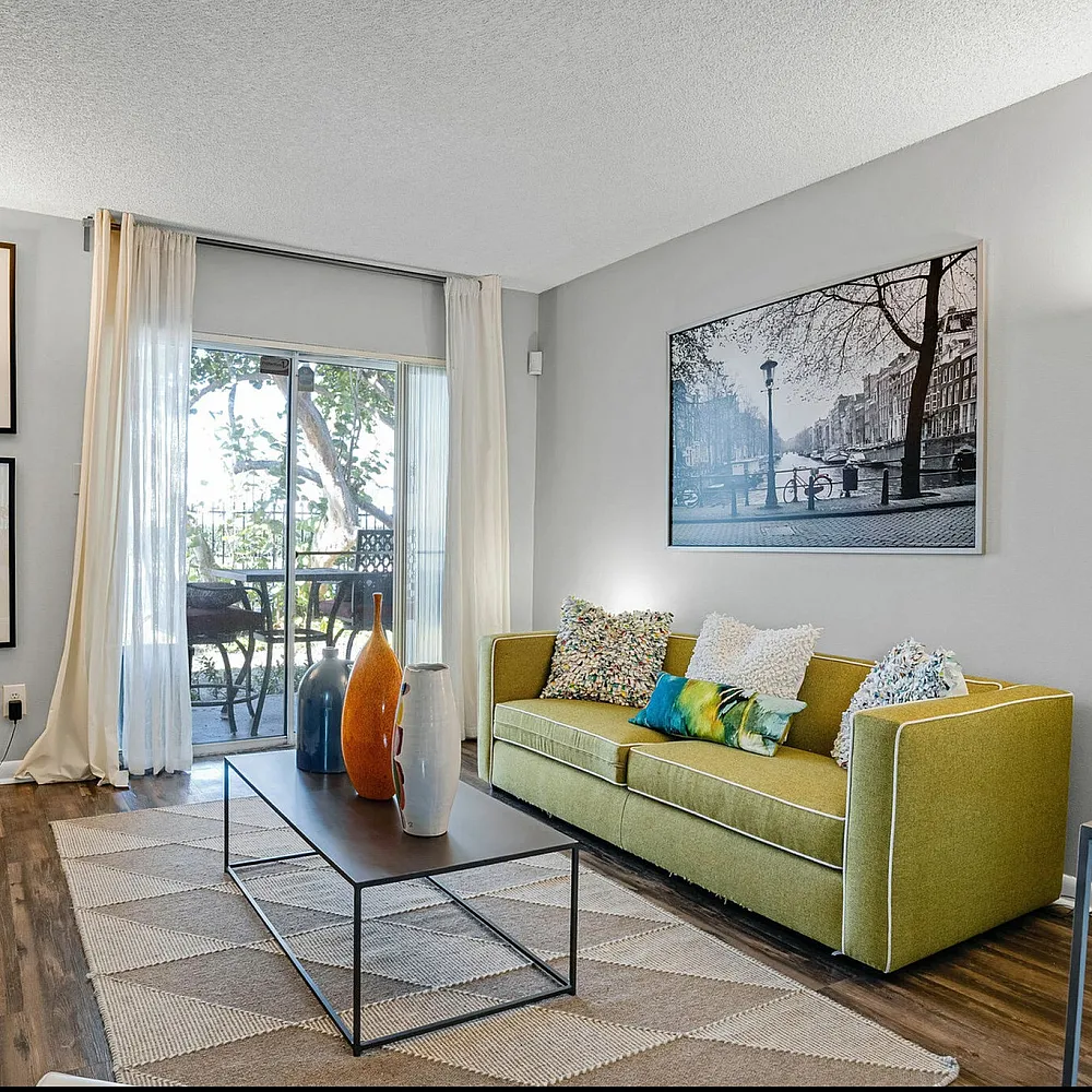 Charming living room with a green sofa, vibrant cushions, and modern art decorating the walls. Large window lets in natural light, illuminating the cozy space with a view of the outdoor patio.