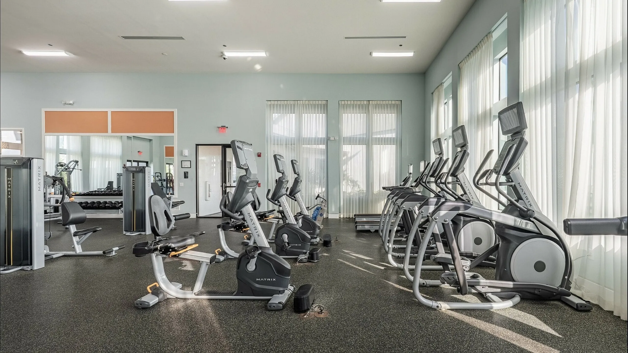 Modern fitness center with top-of-the-line equipment, including treadmills and elliptical trainers, flooded with natural light through large windows.