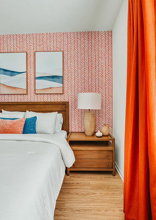 Chic bedroom with vibrant red patterned wallpaper, elegant wooden nightstand, and cozy bedding adorned with colorful pillows.