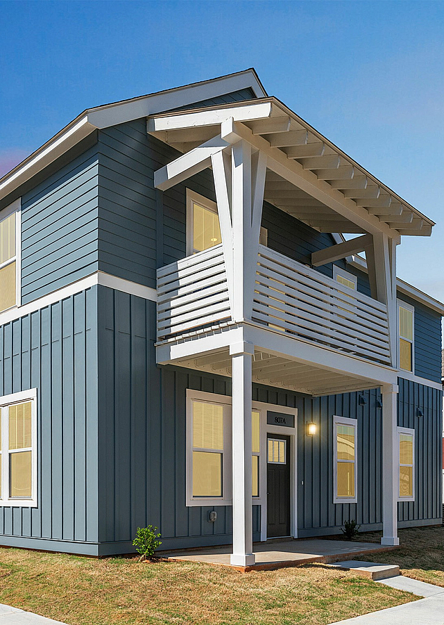 Charming modern two-story blue house with a spacious balcony, large windows, and a beautifully landscaped front yard, set under a clear sky.