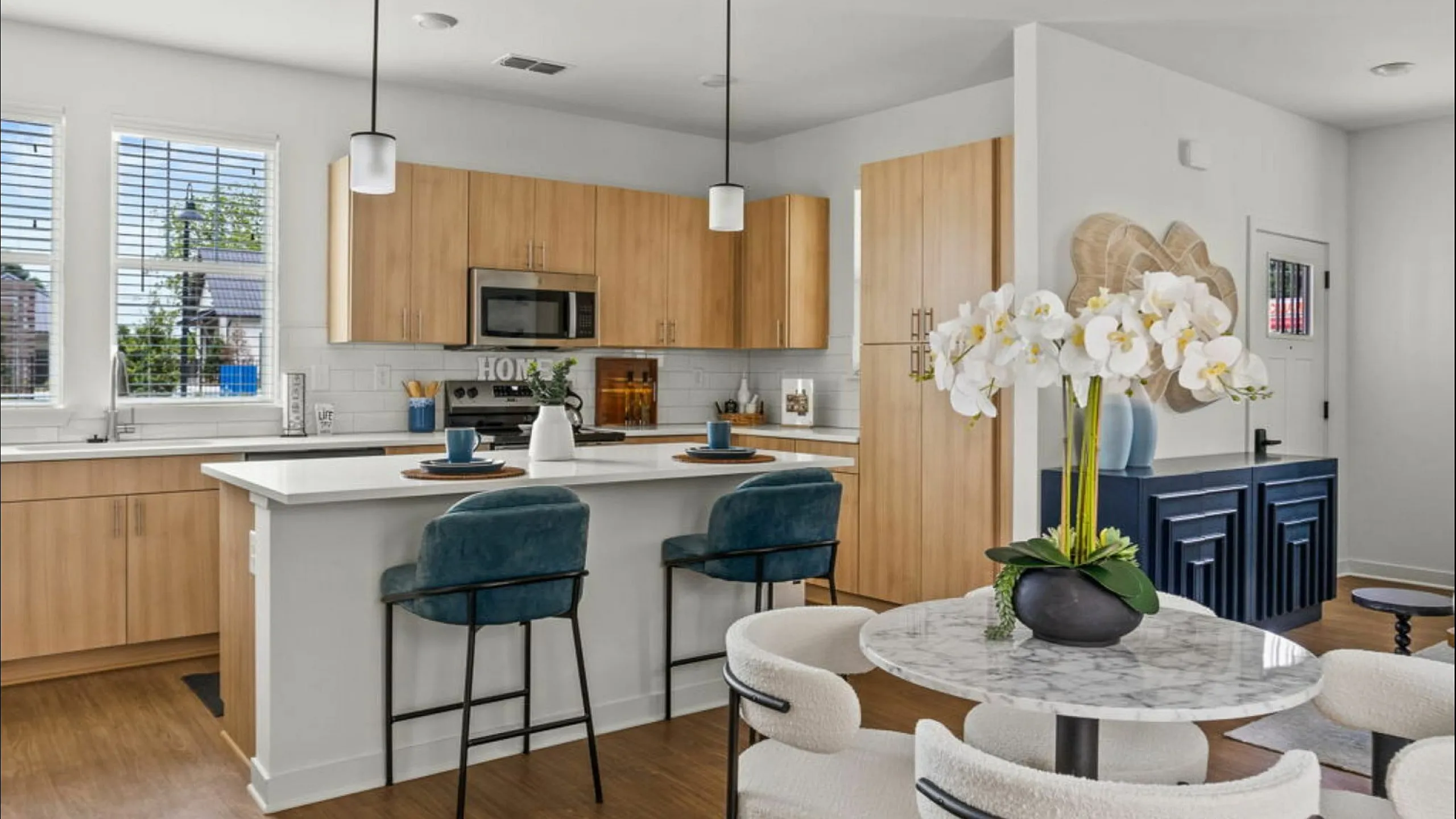 Modern kitchen and dining area with sleek wooden cabinets, white countertops, and elegant lighting. Includes a chic marble table with plush chairs, and lush orchid centerpiece for a luxurious touch.