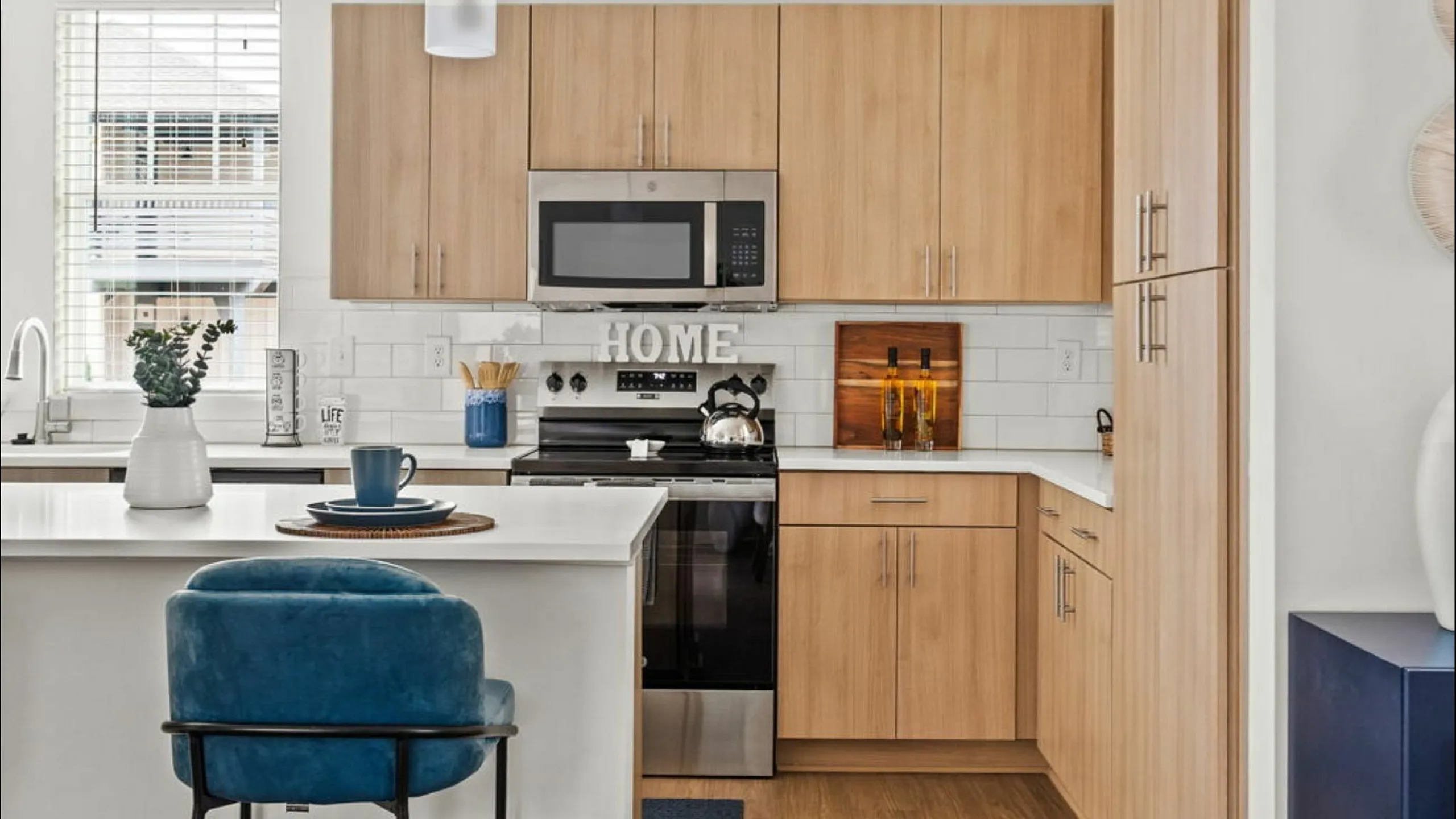 Modern kitchen with sleek wooden cabinets, stylish white countertops, and stainless steel appliances. Charming decor features and a cozy blue chair accentuate the inviting atmosphere.