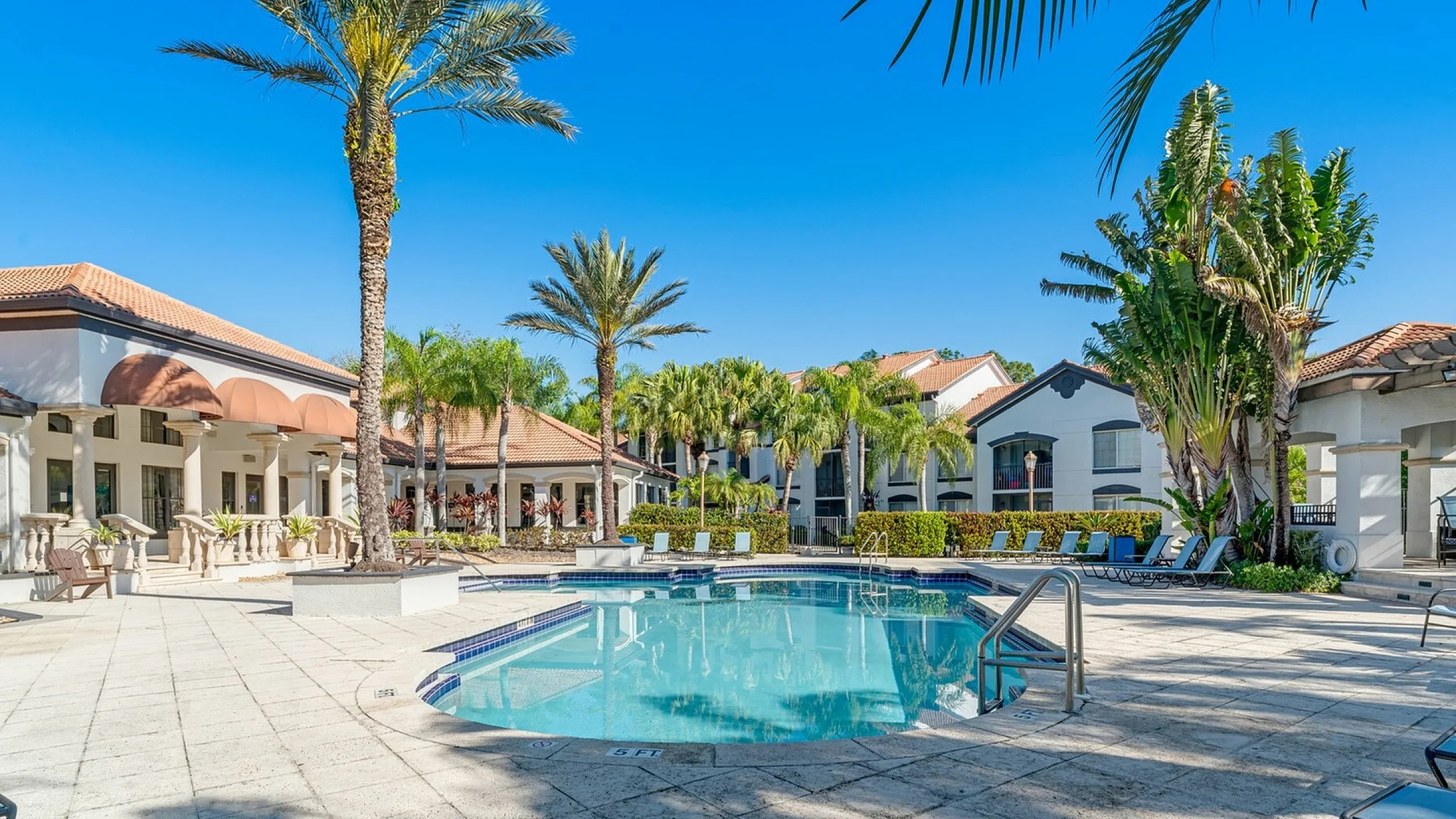 Luxurious apartment complex featuring a sunlit pool area surrounded by palm trees and elegant architecture, perfect for resort-style living.
