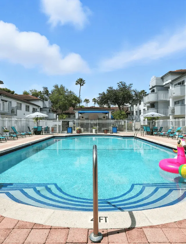 Inviting outdoor pool area surrounded by lush greenery and modern apartments, featuring comfortable lounge chairs and a vibrant pink pool float for relaxation.