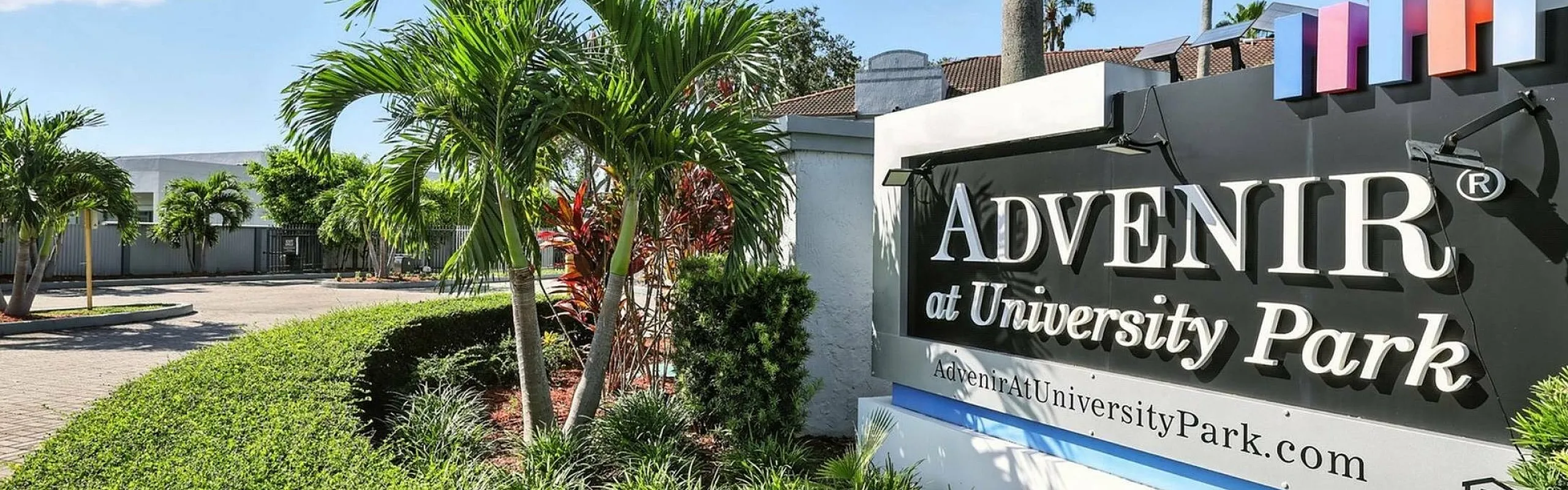 Inviting entrance to Advenir at University Park, featuring lush greenery and palm trees under a bright blue sky.