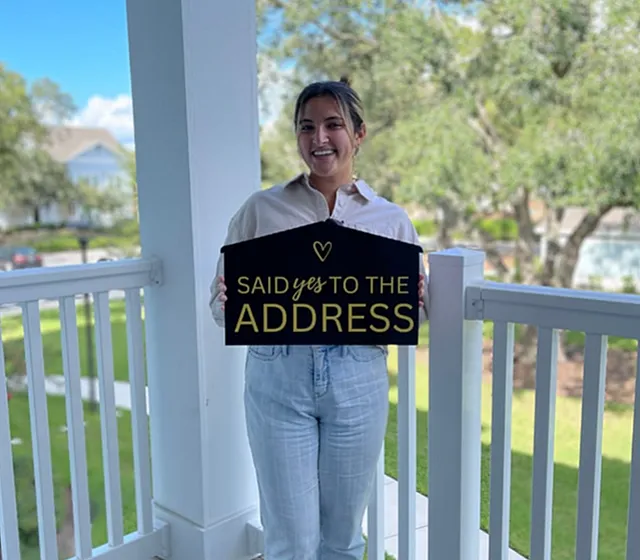 Happy new homeowner on a sunny porch holding a sign that reads 'Said Yes to the Address,' celebrating their picturesque new property.
