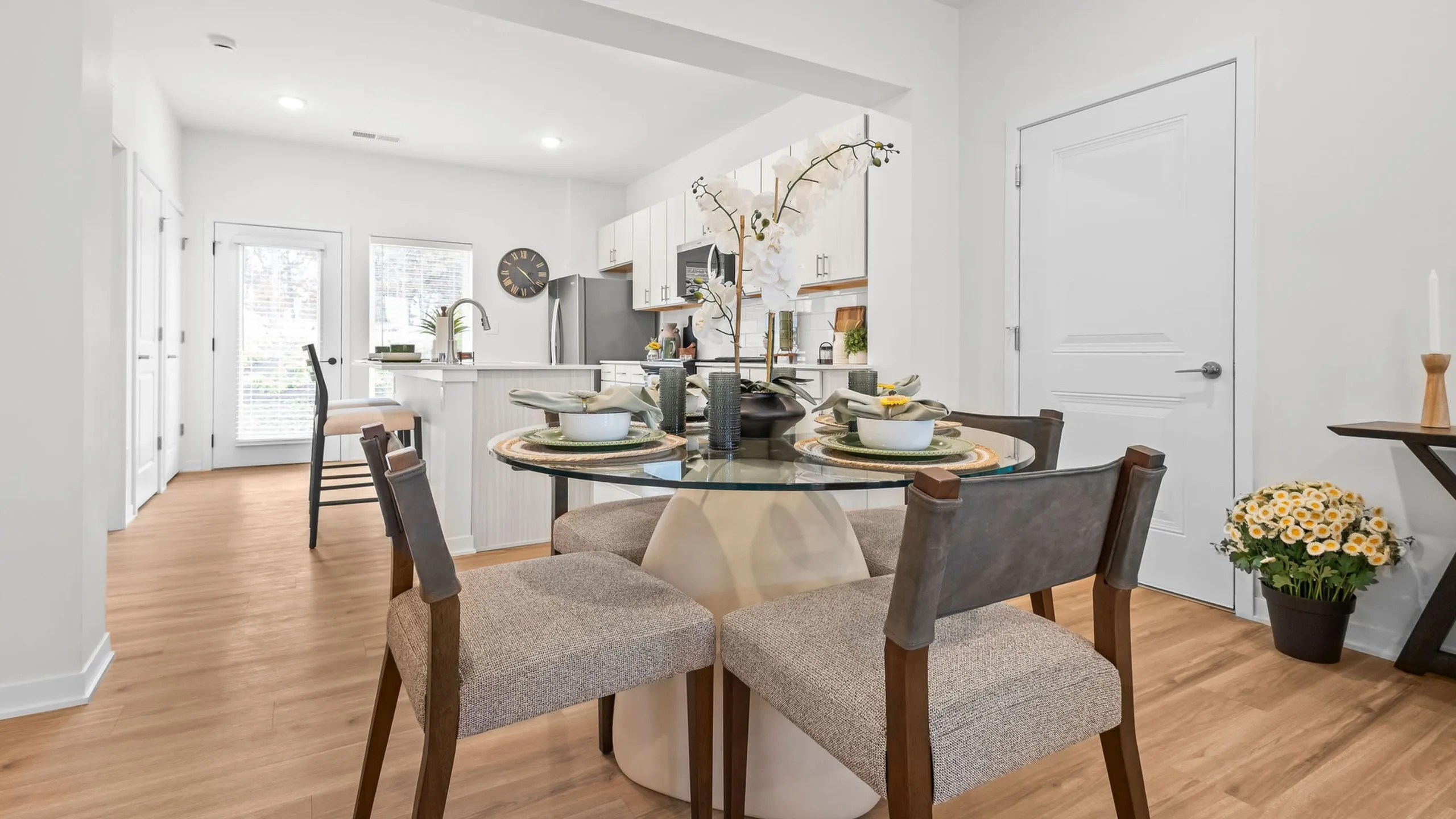 Chic dining area with a modern glass table set for four, adjacent to a sleek, contemporary white kitchen featuring stainless steel appliances and bright natural light.
