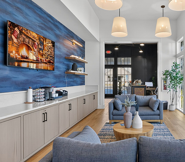 Inviting leasing office room view of the coffee area with modern decor, featuring cozy gray sofas, elegant lighting, and large windows offering ample natural light.