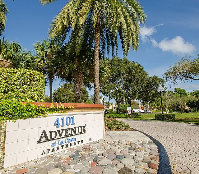 Inviting entryway to Advenir at La Costa Apartments, showcasing lush greenery, palm trees, and a welcoming path under a clear blue sky.