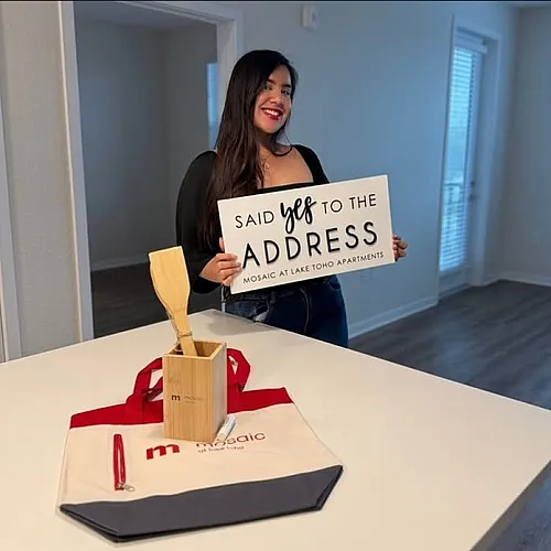 Delighted woman holding a sign that reads 'Said Yes To The Address' at Mosaic at Lake Toho Apartments, showcasing a modern, welcoming interior with natural light and stylish finishes.