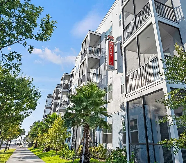 Modern apartment building exterior featuring balconies, lush greenery, and a vibrant tree-lined sidewalk under a clear blue sky.