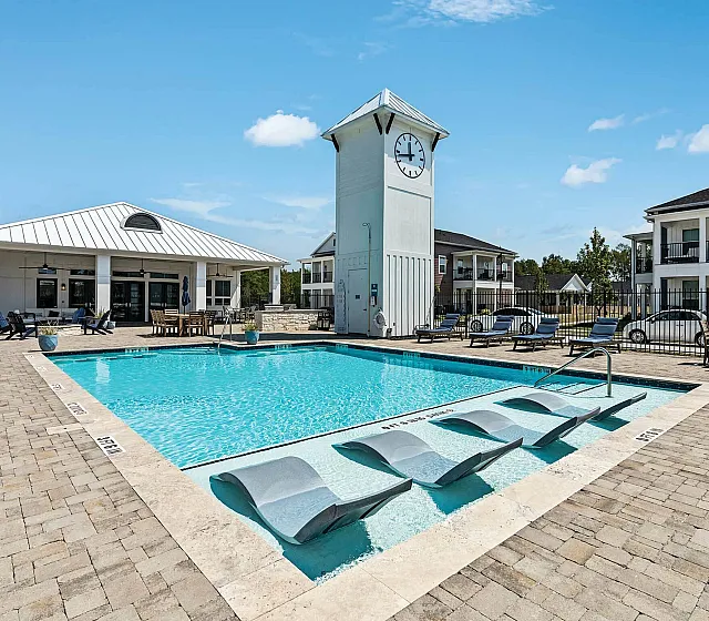Luxurious apartment complex featuring a sparkling swimming pool with sun loungers, a modern clock tower, and stylish outdoor seating under a blue sky.