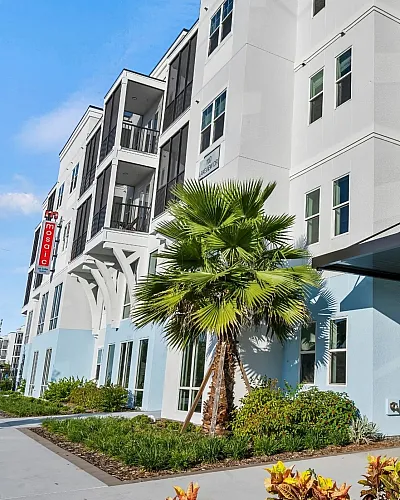 Modern apartment building exterior with sleek balconies, vibrant landscaping, and palm trees under a clear blue sky.