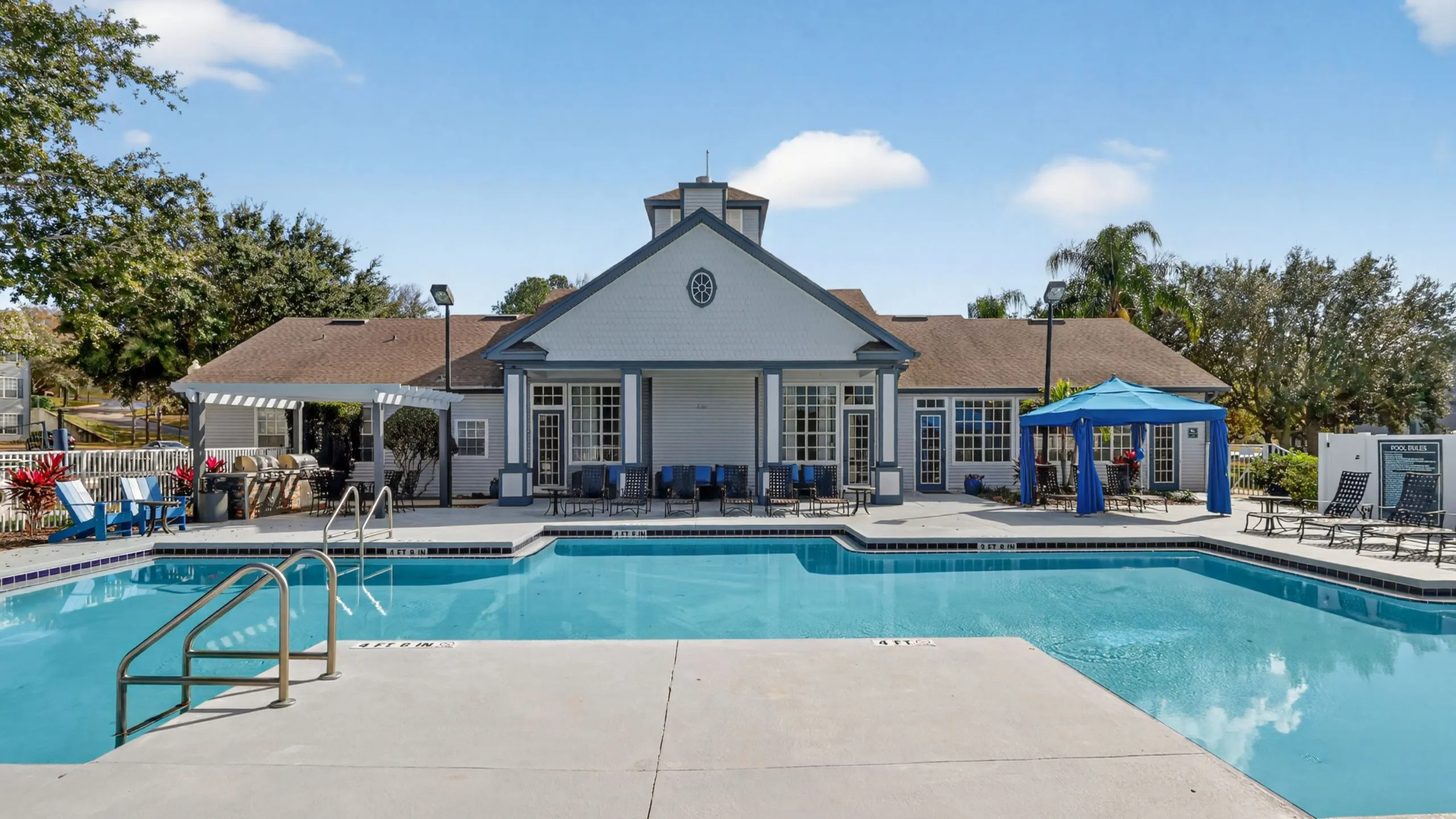 Beautiful residential community with a sparkling blue swimming pool, surrounded by comfortable lounge chairs, and a stylish clubhouse, set against a backdrop of lush greenery and clear blue skies.