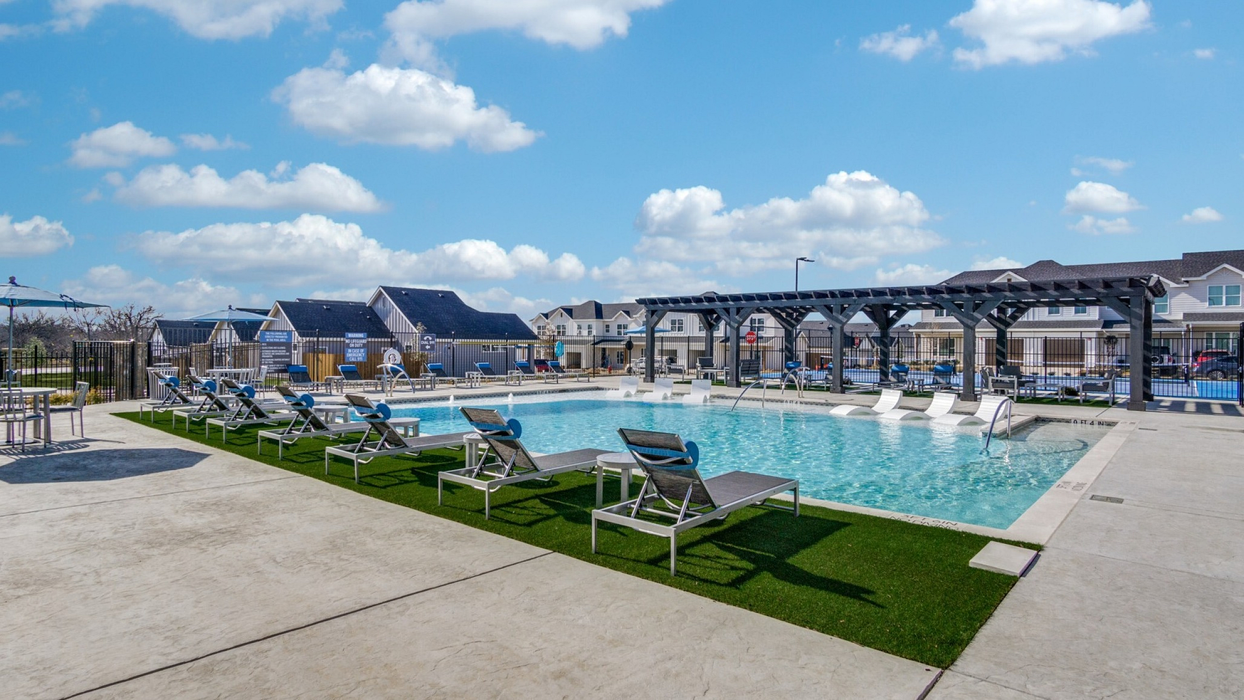 Luxurious outdoor pool area with stylish sun loungers and shaded pergolas under a bright blue sky, surrounded by charming residential homes.